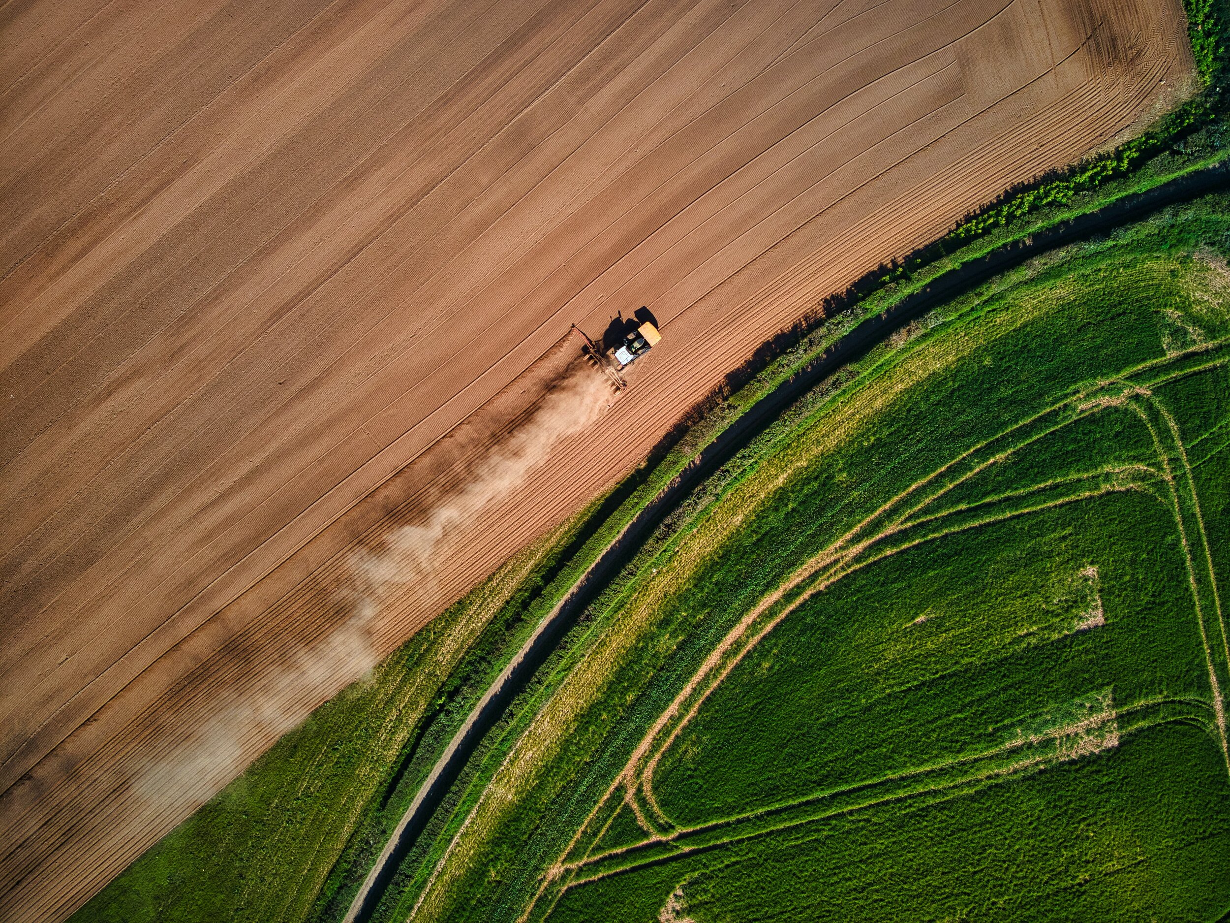 Image description: An aerial photo of a plow trailing dust through a dry field in stark contrast to the bright green pasture on the plot of land beside it.